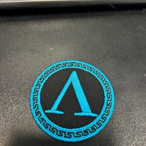 Peut inclure: Écusson rond noir avec un symbole grec Lambda bleu clair au centre. Le symbole est entouré d'une bordure grecque bleu clair. Il s'agit probablement d'un écusson brodé.