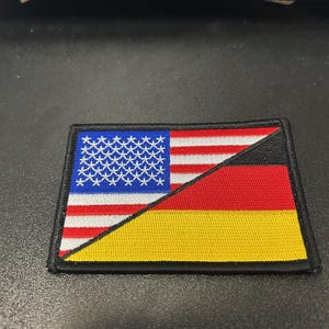 May include: A black patch with the American flag and the German flag sewn together. The flags are sewn together at a diagonal angle, with the American flag on top and the German flag on the bottom.