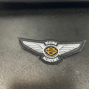 May include: A black and white Harley Davidson Riding Academy patch with a yellow and black Harley Davidson logo and the words "Riding Academy" in white.