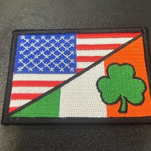 May include: A black patch with an American flag and an Irish flag sewn together. The American flag is on the top and the Irish flag is on the bottom. The flags are sewn together at a diagonal angle. The American flag has red and white stripes and blue stars. The Irish flag has green, white, and orange stripes with a shamrock in the center.