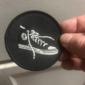 Peut inclure: Un écusson rond noir avec une broderie blanche représentant une basket montante. La chaussure a des lacets blancs et un logo étoile. L'écusson est destiné aux vêtements ou accessoires.