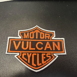 Può includere: Toppa ricamata arancione e nera con il testo "MOTOR VULCAN CYCLES" a forma di scudo.