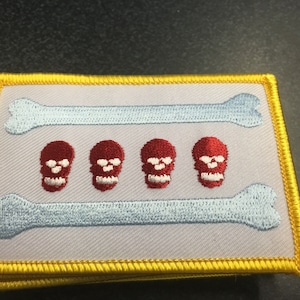 May include: A white embroidered patch with a yellow border. The patch features two blue bones and four red skulls.