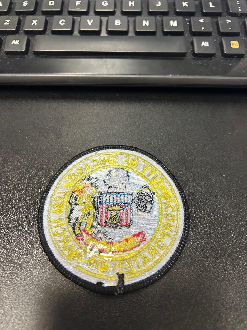May include: A white and yellow embroidered patch with a black border. The patch features a shield with a red, white, and blue design and the text "CHICAGO POLICE DEPARTMENT" around the shield.