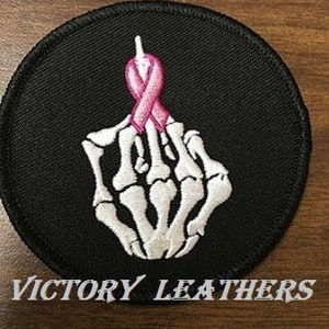 May include: Black embroidered patch with a white skeleton hand giving the middle finger. A pink ribbon is tied around the middle finger.