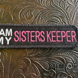May include: A rectangular black embroidered patch with a stitched border. The text "I AM MY" is embroidered in white thread on the left side, and the text "SISTERS KEEPER" is embroidered in pink thread on the right side of the patch.