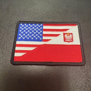 Może przedstawiać: Prostokątna, haftowana naszywka łącząca flagi amerykańską i polską. Amerykańska strona przedstawia niebieski z białymi gwiazdami oraz czerwono-białe paski. Polska strona przedstawia czerwień i biel z białym godłem orła.