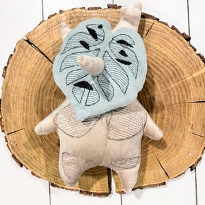 May include: A beige plush toy with a stitched monstera leaf design on its head. The toy is sitting on a wooden surface.