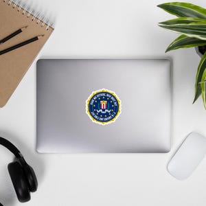 Könnte beinhalten: Ein blauer und goldener Aufkleber mit dem Siegel des Federal Bureau of Investigation (FBI). Das Siegel zeigt einen Schild mit einem Schlüssel, einem Band und den Worten "Treue, Tapferkeit, Integrität". Der Text um das Siegel lautet "The Federal Bureau of Investigation, United States Department of Justice".