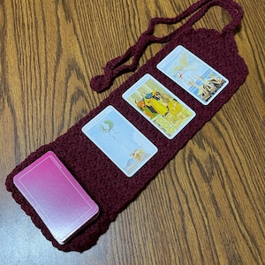 May include: A deep red, crocheted tarot card holder with a loop handle. The holder displays a deck of cards and three tarot cards. The tarot cards are visible on the holder. The holder is on a wooden surface.