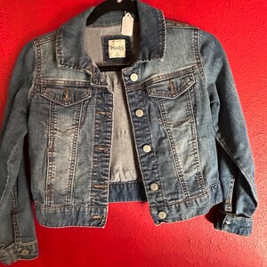 May include: A blue denim jacket with a classic collar, button-up front, and two chest pockets. The jacket is hanging on a black hanger against a red background. The brand label "Mudd" is visible.