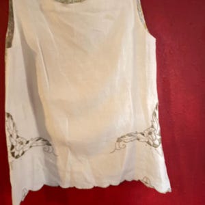 May include: A sleeveless, off-white top with a scalloped hem and decorative embroidery. The top has a floral patterned lining visible at the neckline and shoulder. The garment is hanging against a red background.