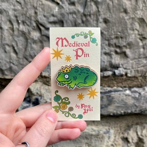 May include: A Medieval Pin featuring a green cartoon lizard wearing a gold crown. The pin is on a cream-coloured card with the words "Medieval Pin" and the artist's name. Decorative stars and floral elements surround the text.