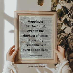 May include: A framed print displaying a quote by Albus Dumbledore: "Happiness can be found, even in the darkest of times, if one only remembers to turn on the light." The text is printed on a vintage dictionary page, within a gold-coloured frame.