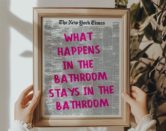 Op de afbeelding: Een ingelijste print met de tekst "WHAT HAPPENS IN THE BATHROOM STAYS IN THE BATHROOM" in felroze letters. De achtergrond is een vintage krantenpagina. De lijst is licht van kleur.
