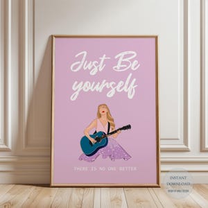 Pode incluir: Uma impressão de arte emoldurada com um fundo roxo claro. A impressão apresenta uma ilustração de uma pessoa tocando uma guitarra azul, com as palavras "Just Be yourself" e "There Is No One Better" em escrita branca. A pessoa está vestindo um vestido rosa.