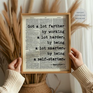 May include: A framed print with a light wood frame. The print features text on a vintage dictionary page that reads, "Got a lot farther by working a lot harder, by being a lot smarter, by being a self-starter." The author is listed as Hamilton.