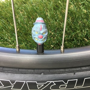 May include: A blue and red clown face bike valve cap with a black stem. The valve cap is attached to a bicycle wheel.