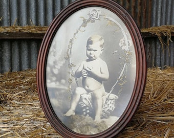 May include: A vintage oval-shaped picture frame with a dark wood finish. The frame contains a black and white photograph of a young child sitting in a basket. The child is wearing no clothes and has a serious expression on their face.