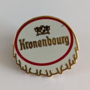May include: A round pin featuring the Kronenbourg logo. The pin has a white center with the brand name in gold lettering, a red border, and a gold-colored edge. The design resembles a bottle cap.