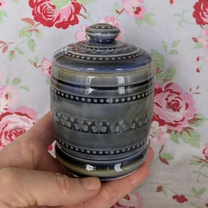 Könnte beinhalten: Ein kleines, blaues Keramikgefäß mit Deckel. Das Gefäß hat eine zylindrische Form mit dekorativen Bändern aus Punkten und floralen Mustern. Der Deckel ist ebenfalls mit einem ähnlichen Muster verziert. Das Gefäß wird vor einem floralen Hintergrund gehalten.