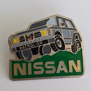 Peut inclure: Épinglette métallique Nissan Patrol GR. L'épinglette représente un SUV gris avec des pneus noirs et des phares jaunes, sur un fond vert avec le mot "NISSAN" en lettres dorées. L'épinglette est un objet de collection.