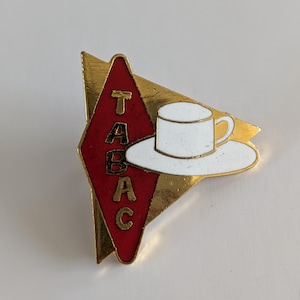 May include: A vintage pin badge with a red diamond displaying the word "TABAC" in gold lettering. A white teacup and saucer are on the right side. The pin has a gold border and a gold triangle shape.