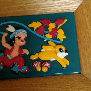 May include: A framed ceramic tile depicting a colourful scene of a girl in a red dress and a yellow rabbit running through a forest. The tile has a teal background with red and yellow leaves.