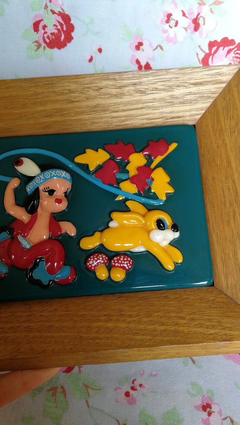 May include: A framed ceramic tile depicting a colourful scene of a girl in a red dress and a yellow rabbit running through a forest. The tile has a teal background with red and yellow leaves.