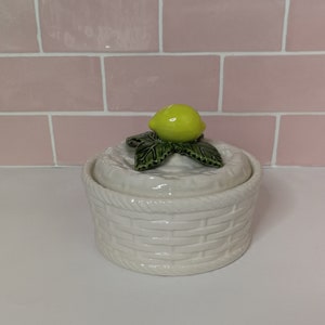 Puede incluir: Jarrón de cerámica blanca con diseño de cesta tejida. La tapa presenta una hoja verde y un limón amarillo.
