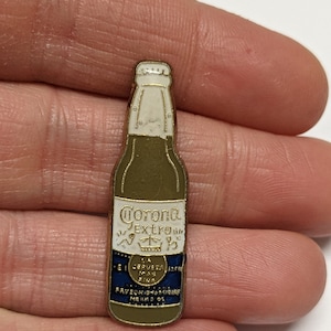 Puede incluir: Pin de metal dorado con forma de botella de cerveza Corona Extra. La botella tiene una etiqueta blanca con el texto "Corona Extra" y un logotipo de corona. La etiqueta también tiene el texto "La Cerveza Mas Fina" y "FAVORECIDA POR MILLONES DE.