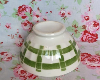 May include: A white ceramic bowl with a green and white checkered pattern. The bowl is sitting on a floral fabric background.