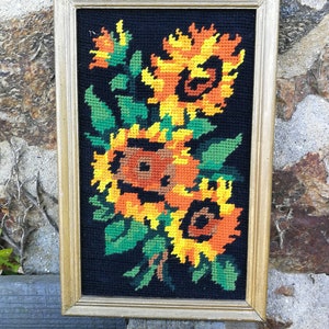 May include: A framed needlepoint embroidery featuring a bouquet of sunflowers in shades of yellow, orange, and brown against a black background. The frame is gold.