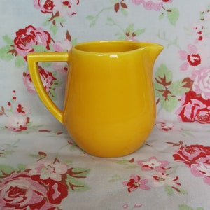 May include: A bright yellow ceramic creamer pitcher with a rounded body and a handle. The pitcher is sitting on a floral tablecloth.