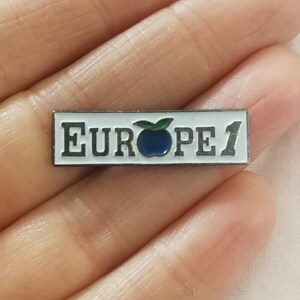 Op de afbeelding: Een witte emaille pin met een blauwe appel en de tekst "EUROPE 1" in zwart.