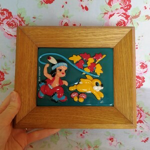 May include: A framed 3D artwork featuring a cartoon Native American girl in a red and blue outfit holding a lasso, a yellow rabbit, and red and green leaves on a teal background. The artwork is signed "Guy Cochet".