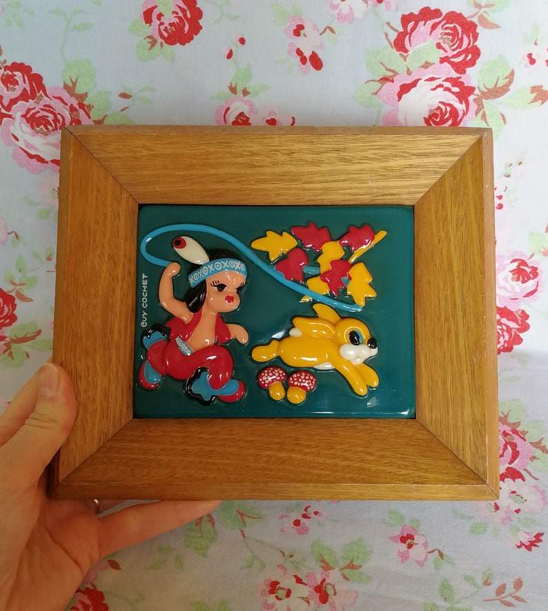 May include: A framed 3D artwork featuring a cartoon Native American girl in a red and blue outfit holding a lasso, a yellow rabbit, and red and green leaves on a teal background. The artwork is signed "Guy Cochet".