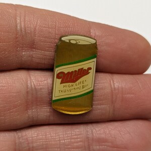 May include: A gold-coloured metal pin shaped like a can of Miller High Life beer. The can has a red and white label with the words "Miller High Life" and "The Champagne of Beers".