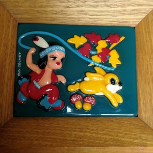May include: A framed ceramic wall art piece featuring a cartoon girl in a red and blue outfit holding a lasso, a yellow rabbit, and red and yellow mushrooms on a teal background. The frame is made of wood and has a brown finish. The text "GUY COCHET" is visible on the frame.