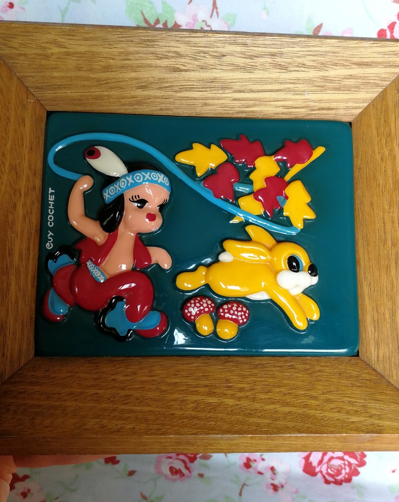 May include: A framed ceramic wall art piece featuring a cartoon girl in a red and blue outfit holding a lasso, a yellow rabbit, and red and yellow mushrooms on a teal background. The frame is made of wood and has a brown finish. The text "GUY COCHET" is visible on the frame.