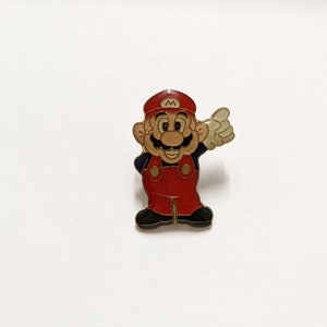 Könnte beinhalten: Eine Emaille-Anstecknadel mit einer Comicfigur von Mario aus der Super Mario Bros. Videospielserie. Mario trägt seine charakteristischen roten Latzhosen und eine Mütze mit einem weißen 'M' darauf. Er lächelt und winkt mit der rechten Hand.