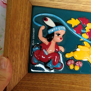 May include: A framed ceramic tile featuring a colourful illustration of a girl in a red and blue outfit holding a lasso. The tile is set against a teal background with yellow and red leaves. The tile is signed "Guy Cochet".
