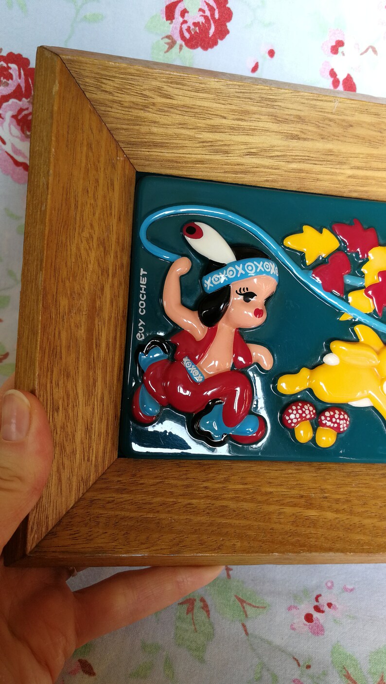 May include: A framed ceramic tile featuring a colourful illustration of a girl in a red and blue outfit holding a lasso. The tile is set against a teal background with yellow and red leaves. The tile is signed "Guy Cochet".