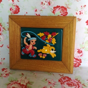 May include: A framed ceramic tile featuring a colourful illustration of a Native American girl in a red dress and a blue headdress, holding a lasso and riding a yellow rabbit. The tile has a green background with red, yellow, and orange leaves. The tile is framed in a brown wooden frame.