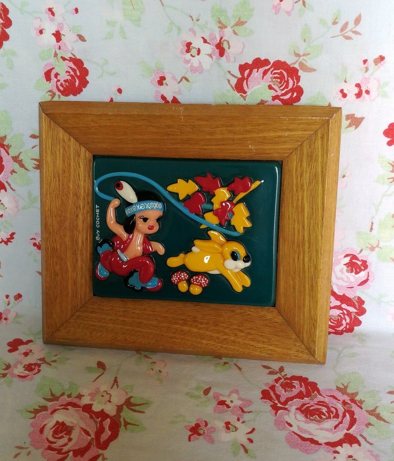 May include: A framed ceramic tile featuring a colourful illustration of a Native American girl in a red dress and a blue headdress, holding a lasso and riding a yellow rabbit. The tile has a green background with red, yellow, and orange leaves. The tile is framed in a brown wooden frame.