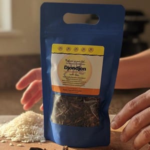 May include: A blue pouch of DjonDjon mushrooms with a clear window, alongside a pile of white rice, dried mushrooms, and bay leaves on a wooden chopping board. The pouch has a yellow and orange label with text. The scene is lit by sunlight.