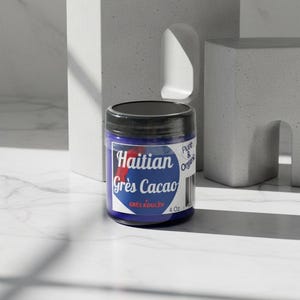 Op de afbeelding: Een blauwe pot met een zwart deksel, met het opschrift "Haitian Gr&egrave;s Cacao" met een rode accent. De pot bevat 113g product en staat tegen een minimalistische, getextureerde achtergrond.