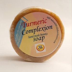 May include: A round, unwrapped bar of turmeric complexion soap. The soap is a light brown color and has a label with the words "Turmeric Complexion soap" in red and blue. The label also includes the words "hand made beauty" and a logo.