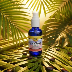 May include: A blue glass bottle with a white pump dispenser, labeled "Palma Christi Oil", sits on green palm leaves and bamboo stalks. Brown seeds are scattered around the bottle. The background features more green palm leaves.