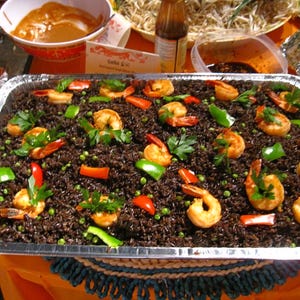 May include: A large rectangular foil tray of black rice, prawns, and vegetables. The prawns are cooked and arranged on top of the rice, with red and green peppers, peas, and parsley. Other dishes are visible in the background.